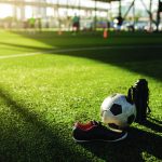Football, cool flask and black red sports shoes on green artificial turf with blurry kid soccer player training. Sunshine and shadows of soccer training equipment.